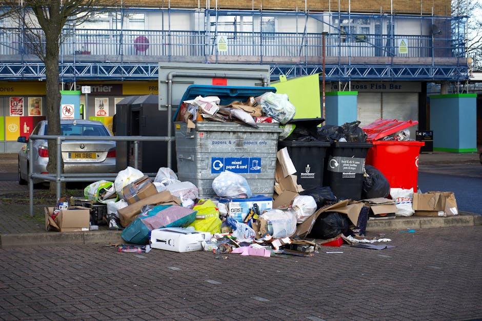 A large assortment of mixed rubbish and waste materials overflowing from a central grey recycling bin labeled for paper and cardboard, situated on a paved area in an urban environment. The waste inclu